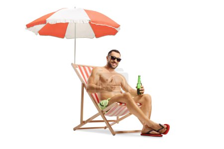 Photo for Young man in swimwear sitting on a beach chair with a bottle of bear under an umbrella isolated on white background - Royalty Free Image