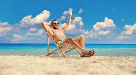 Photo for Young man in swimwear sitting on a beach chair by the sea and waving - Royalty Free Image