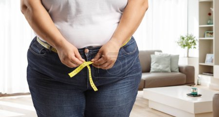 Foto de Mujer con sobrepeso que usa jeans y mide la cintura dentro de una sala de estar - Imagen libre de derechos