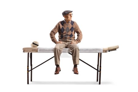 Foto de Hombre mayor sentado en una cama de fisioterapia y mirando al lado aislado sobre fondo blanco - Imagen libre de derechos