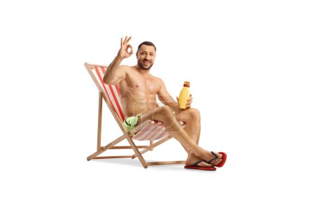 Photo for Young man sitting on a beach chair holding a bottle of sunscreen and gesturing ok sign isolated on white background - Royalty Free Image