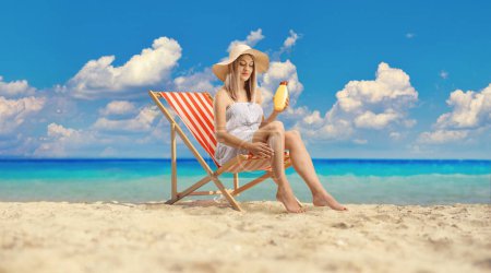 Photo for Woman sitting on a deck chair and applying sun cream on legs by the sea - Royalty Free Image