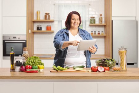 Téléchargez les photos : Femme avec une tablette numérique à la recherche d'une recette en ligne et debout dans une cuisine - en image libre de droit