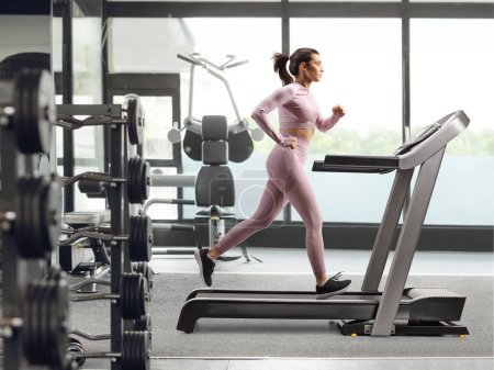 Foto de Foto de perfil completo de una mujer corriendo en una cinta de correr en el gimnasio - Imagen libre de derechos