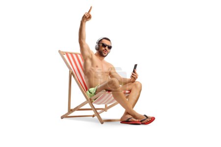Photo for Young man in swimwear sitting on a beach chair and listening to music from a smartphone isolated on white backgroun - Royalty Free Image