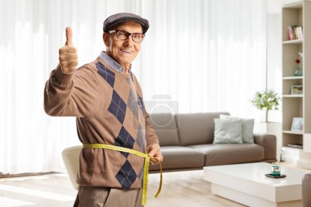 Téléchargez les photos : Homme âgé mesurant la taille avec un ruban adhésif à l'intérieur d'un salon et geste pouces levés - en image libre de droit