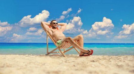 Foto de Hombre joven en traje de baño y gafas de sol sentado en una silla de playa por el se - Imagen libre de derechos