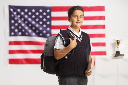 Foto de Chico de uniforme escolar sosteniendo un libro y llevando una mochila delante de una bandera de EE.UU. - Imagen libre de derechos