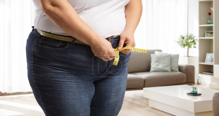 Téléchargez les photos : Femme obèse portant un jean et mesurant la taille dans une pièce - en image libre de droit