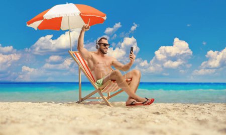 Photo for Young man in swimwear sitting on a beach chair and enjoying with headphones by the sea - Royalty Free Image