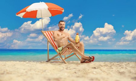 Foto de Hombre joven aplicando crema solar en el pecho sentado en una silla de playa junto al mar - Imagen libre de derechos
