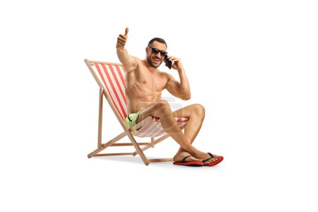 Téléchargez les photos : Jeune homme en maillot de bain assis sur une chaise de plage à l'aide d'un téléphone portable et geste pouces vers le haut isolé sur fond blanc - en image libre de droit