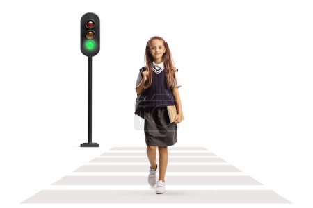 Téléchargez les photos : Portrait complet d'une écolière en uniforme portant un sac à dos et un livre et traversant une rue isolée sur fond blanc - en image libre de droit