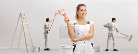 Téléchargez les photos : Joyeux décorateur féminin tenant un pinceau et un seau de couleur et d'autres travailleurs peignant un mur - en image libre de droit