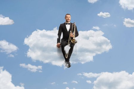 Photo for Male musician in a black suit and bow-tie sitting on a cloud with a trombone - Royalty Free Image