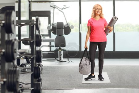 Téléchargez les photos : Portrait complet d'une femme mûre à la salle de gym tenant un tapis d'exercice et un sac de sport - en image libre de droit