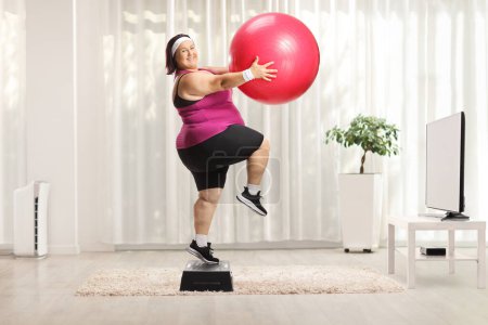 Téléchargez les photos : Femme en surpoids faisant de l'aérobic étape à la maison devant la télévision avec une balle de fitness - en image libre de droit