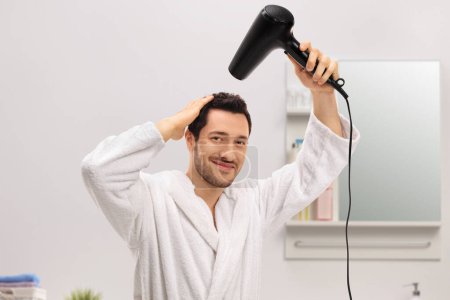 Téléchargez les photos : Le type en peignoir se séchant les cheveux avec un sèche-cheveux dans une salle de bain - en image libre de droit