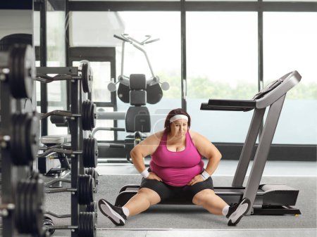 Foto de Mujer cansada con sobrepeso sentada en una cinta de correr en el gimnasio - Imagen libre de derechos