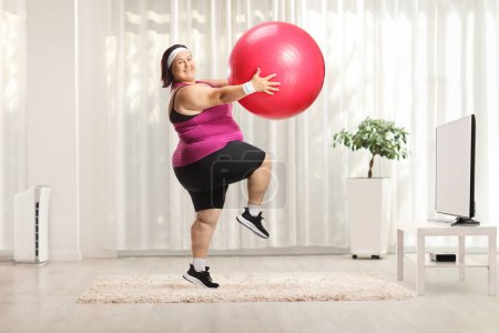 Téléchargez les photos : Femme en surpoids faisant de l'exercice avec une balle de fitness devant une télévision à la maison - en image libre de droit