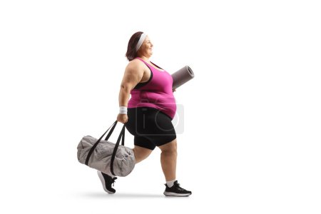 Téléchargez les photos : Jeune femme en surpoids en vêtements de sport portant un sac de sport et marchant isolé sur fond blanc - en image libre de droit