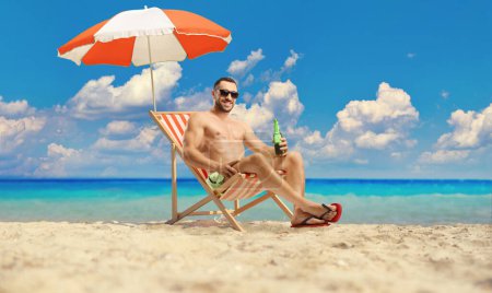 Téléchargez les photos : Homme à la plage au bord de la mer tenant une bouteille de bière et assis sous le parapluie - en image libre de droit