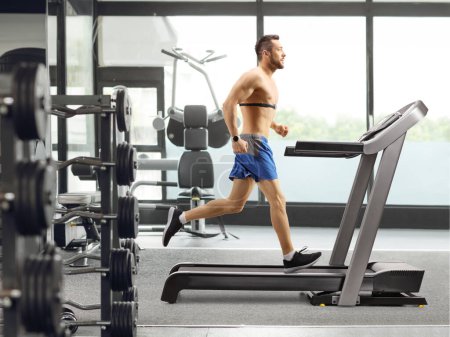 Photo for Full length profile shot of a man in a gym running on a treadmill with a chest monitor - Royalty Free Image