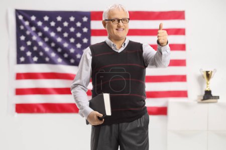 Téléchargez les photos : Professeur masculin tenant un livre et montrant les pouces devant un drapeau américain - en image libre de droit