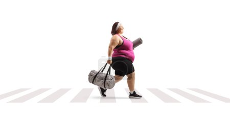 Photo for Overweight young woman in sportswear carrying a bag and walking at a pedestrian crosswalk isolated on white background - Royalty Free Image