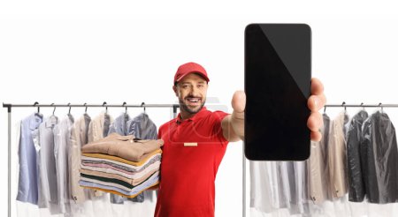 Foto de Trabajador sosteniendo una pila de ropa y mostrando un teléfono inteligente en la tintorería aislado sobre fondo blanco - Imagen libre de derechos