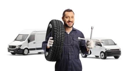 Téléchargez les photos : Mécanicien automobile tenant un pneu sur son épaule et une clé à molette devant des fourgonnettes isolées sur fond blanc - en image libre de droit