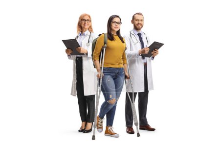 Téléchargez les photos : Étudiante avec des béquilles debout devant des médecins isolés sur fond blanc - en image libre de droit
