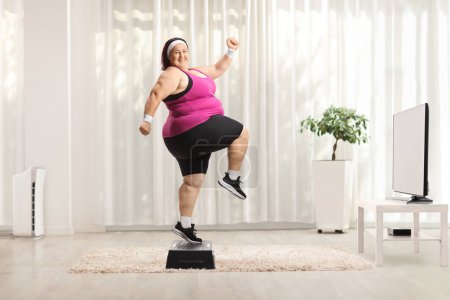 Foto de Alegre mujer con sobrepeso haciendo ejercicio aeróbicos paso en casa delante de una televisión - Imagen libre de derechos