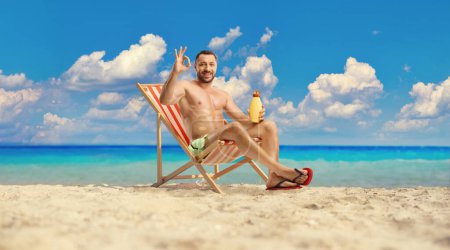 Photo for Man sitting on a deck chair holding a sun lotion and gesturing great at the beach by the sea - Royalty Free Image