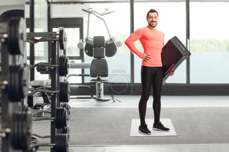 Téléchargez les photos : Portrait complet d'un jeune homme en forme tenant une plate-forme aérobie étape à la salle de gym - en image libre de droit