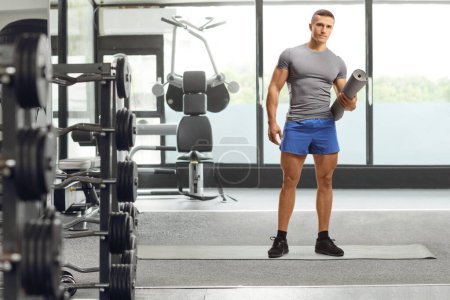 Photo for Full length portrait of a young fit man in sportswear holding an exercise mat inside a gym - Royalty Free Image
