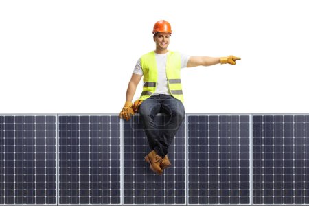 Photo for Construction worker with a helmet sitting on a solar panel and pointing to the side isolated on white background - Royalty Free Image