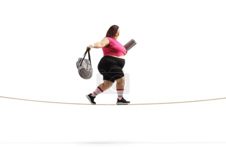 Foto de Foto lateral de una mujer con sobrepeso con una bolsa deportiva y una esterilla de ejercicio caminando sobre una cuerda floja aislada sobre fondo blanco - Imagen libre de derechos