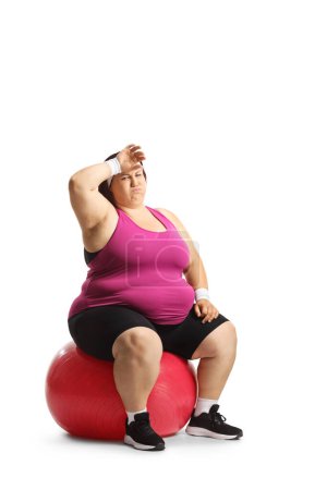 Foto de Mujer cansada y con sobrepeso sentada en una bola de fineza aislada sobre fondo blanco - Imagen libre de derechos