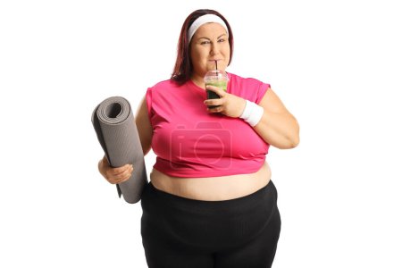 Photo for Smiling overweight woman drinking a healthy green smoothie and holding an exercise mat isolated on white background - Royalty Free Image