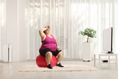 Foto de Cansada mujer con sobrepeso sentada en una pelota fintess delante de la televisión en casa - Imagen libre de derechos