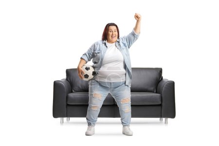 Téléchargez les photos : Jeune femme en surpoids acclamant avec un ballon de football devant un canapé en cuir noir isolé sur fond blanc - en image libre de droit