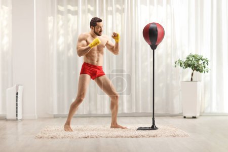 Téléchargez les photos : Jeune homme en short rouge faisant du kickboxing avec un stand de boxe à la maison dans un salon - en image libre de droit