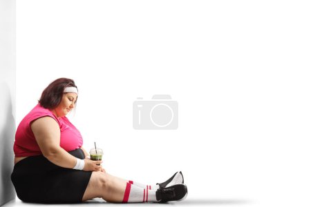 Photo for Sad overweight woman holding a healthy green and sitting on the ground isolated on white background - Royalty Free Image