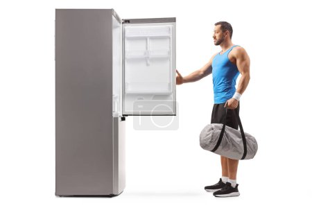 Photo for Fit man holding a sports bag and opening an empty fridge isolated on white background - Royalty Free Image