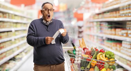 Photo for Shocked mature man with a shopping cart holding a bill at the supermarket - Royalty Free Image