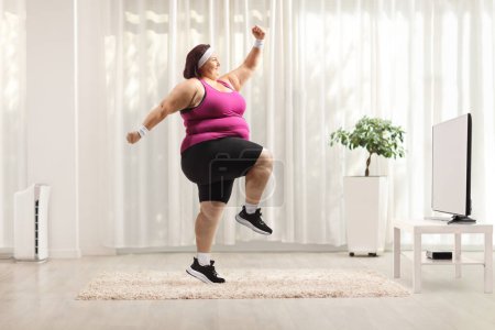 Photo for Full length profile shot of an young overweight woman watching tv and exercising - Royalty Free Image