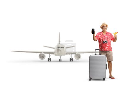 Photo for Mature male tourist with a suitcase holding a cocktail and showing a smartphone in front of an airplane isolated on white background - Royalty Free Image