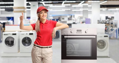 Photo for Female sales asisstant standing next to an electrcal oven inside a shop and gesturing thumbs up - Royalty Free Image