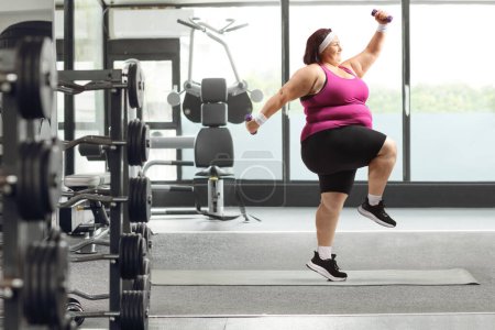 Foto de Foto de perfil completo de una mujer con sobrepeso haciendo ejercicio en un gimnasio y saltando con pesas - Imagen libre de derechos